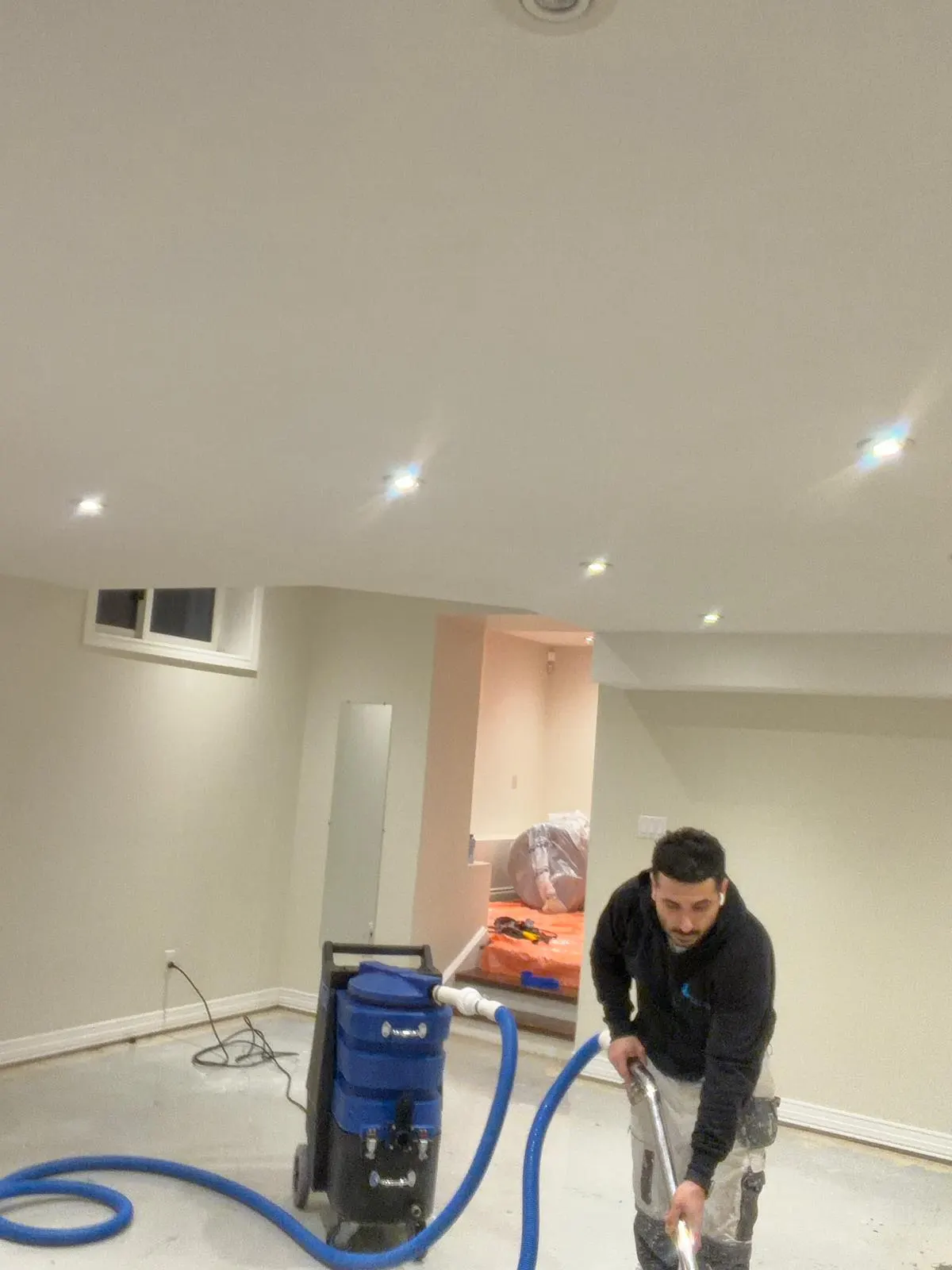 Technician using a hose and portable extraction unit to pull water out of the floor in a finished basement, with furniture wrapped in plastic behind him